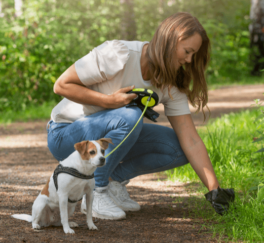Woman kneeling on a path outdoors with a small dog, holding a leash and using a BioBag dog poo bag to pick up waste